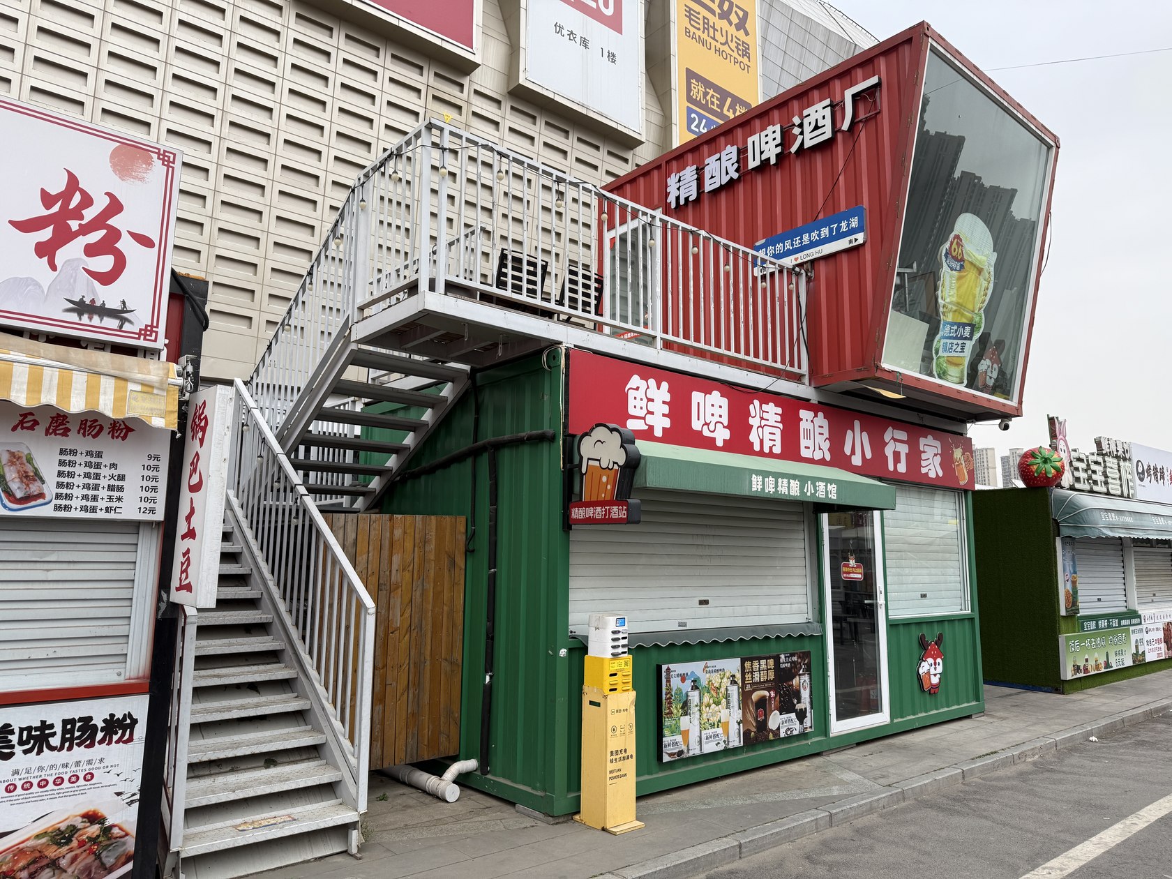 龙湖锦艺城两层集装箱啤酒屋使用阶段照片,展示啤酒销售门头、首层售卖窗口、玻璃门和外置楼梯。